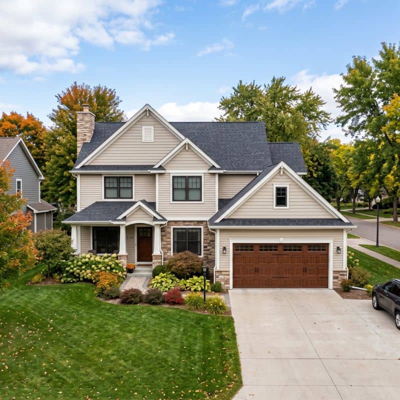 A beautiful two-story home in St. Augusta, MN, with beige siding, stone accents, and a manicured lawn. The high-quality carriage-style dark wood garage door was installed by After Hours Garage Doors.